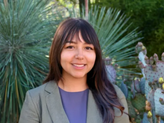 Person wearing a grey blazer and purple shirt, smiling in front of green plants