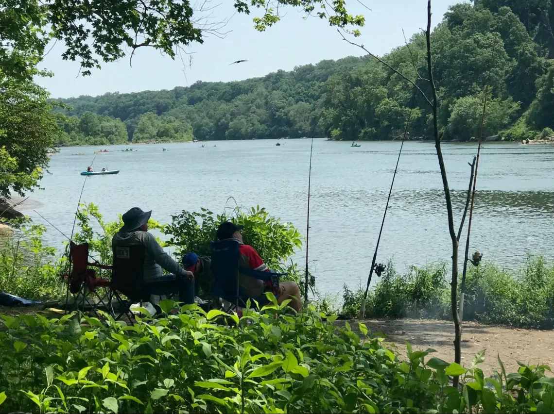 Fisherfolk sitting and fishing at the edge of the river bank, kayakers riding on the water in the distance.