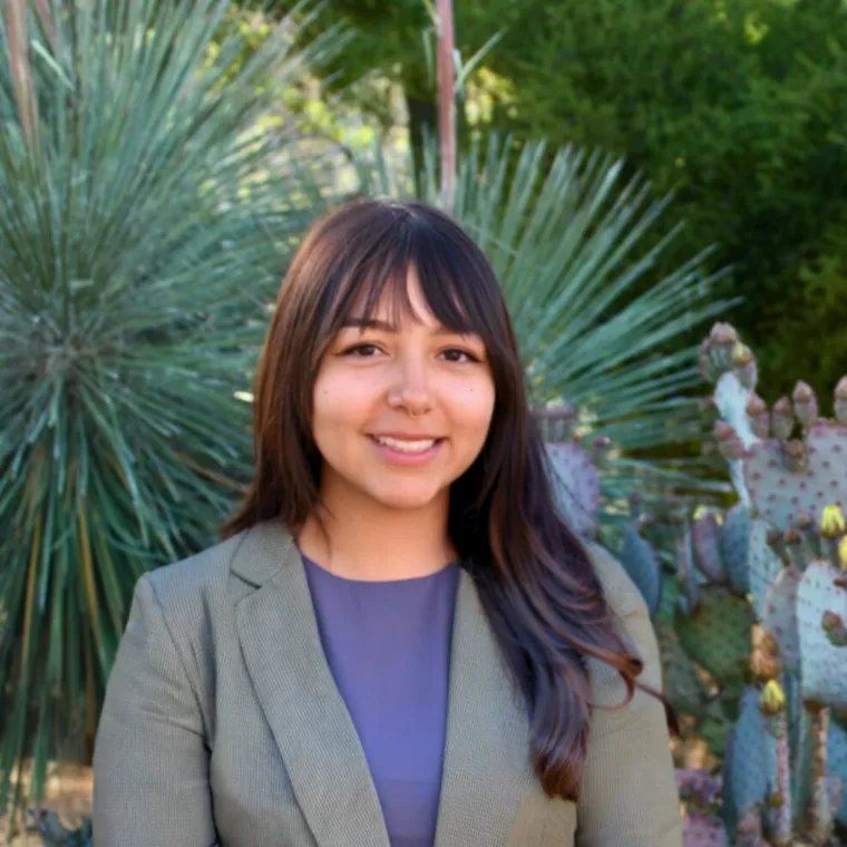 Person wearing a grey blazer and purple shirt, smiling in front of green plants