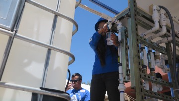 Two people stand near a Solar Nanofiltration (SNF) water device. One person is drinking water from a cup, and the other is filling a cup from the tap on the SNF unit.